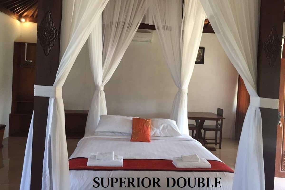 A Superior Double bed with white linens, an orange accent pillow, and a red bedspread. The room has a wooden ceiling and a white canopy over the bed.
