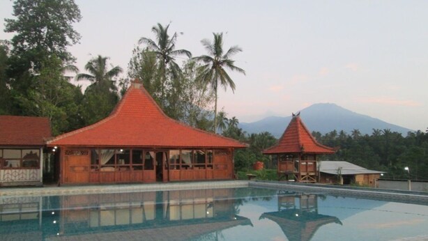 Área de piscina com duas estruturas de madeira, uma com telhado vermelho e outra com telhado cinzento, cercada por palmeiras e uma montanha ao fundo.