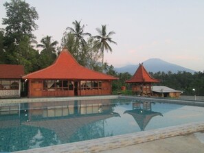 A pool area with two wooden structures, one with a red roof and another with a grey roof, surrounded by palm trees and a mountain in the background. - Kampoeng Joglo Ijen Banyuwangi (Banyuwangi)