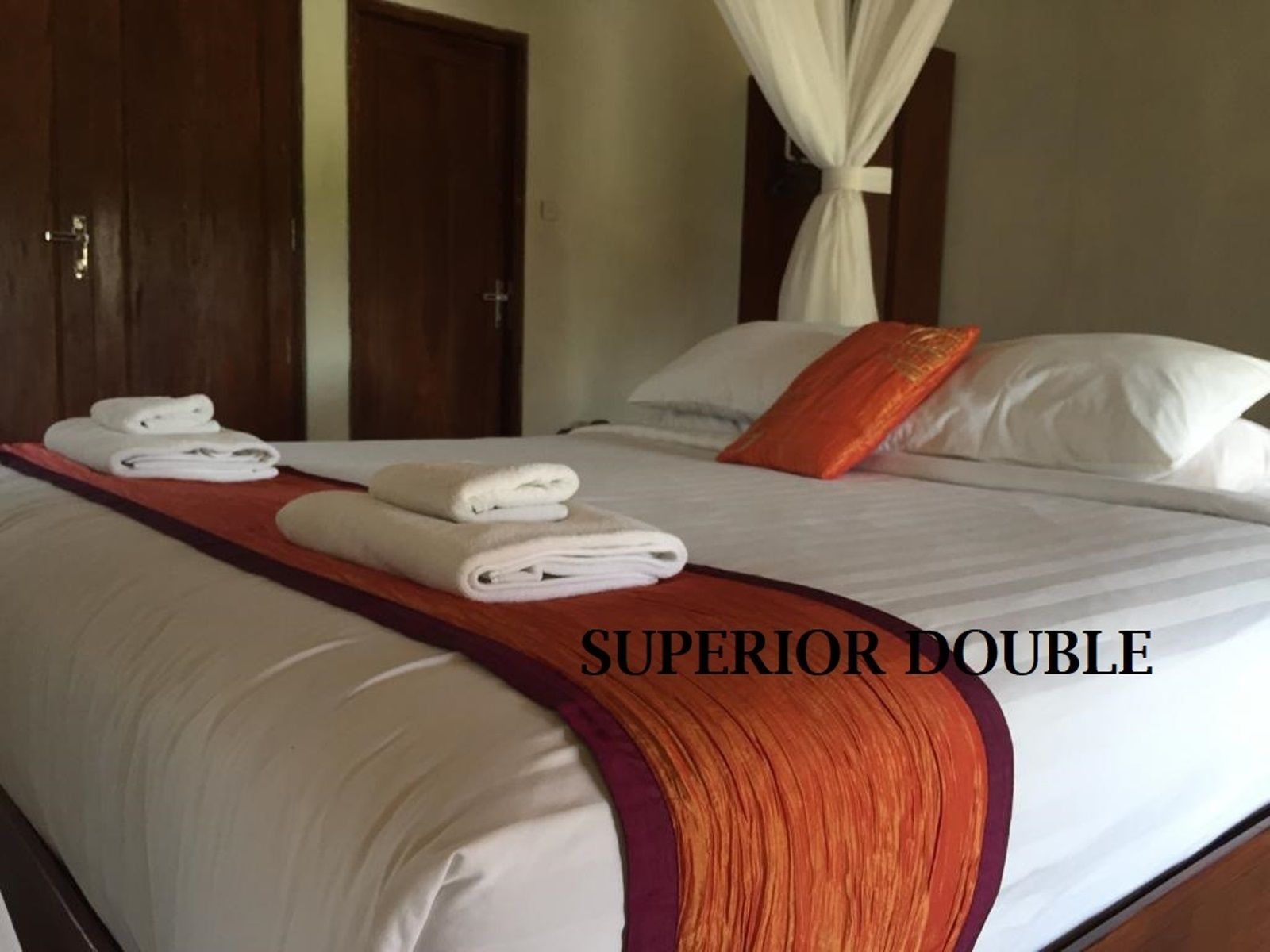 Habitación de hotel con cama doble, ropa de cama blanca, colcha roja y naranja, y dosel.