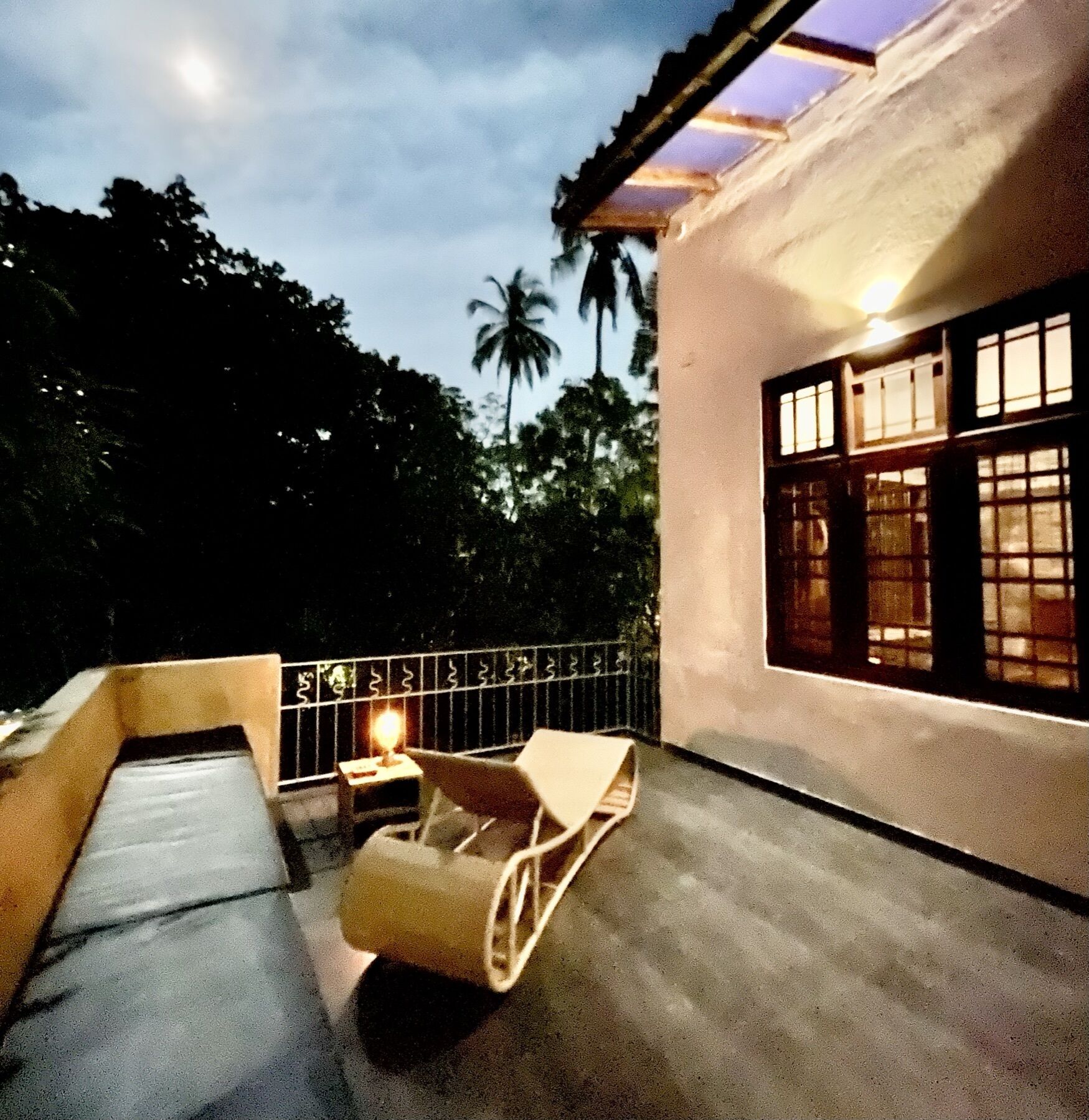 A balcony with a single chair and a table, a window with a view of trees, and a night sky.