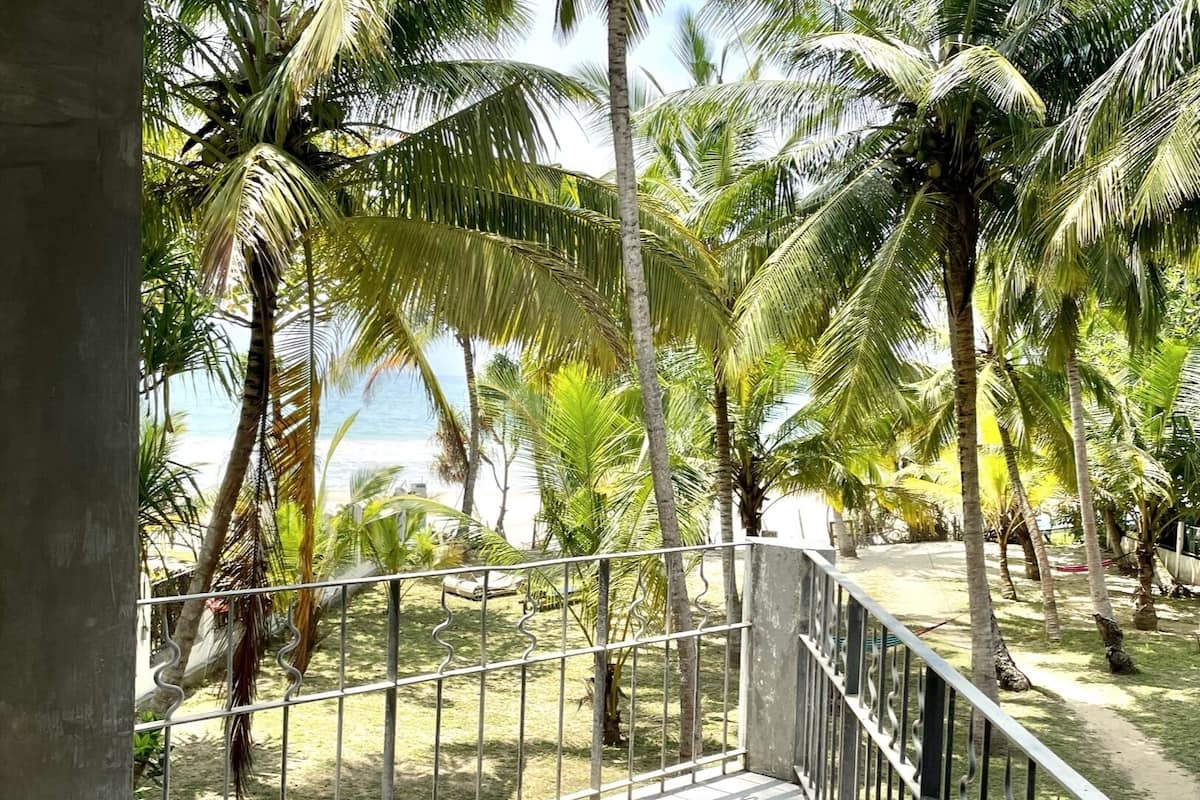 A balcony with tiled flooring and metal railing overlooking a grassy area with palm trees and a body of water.