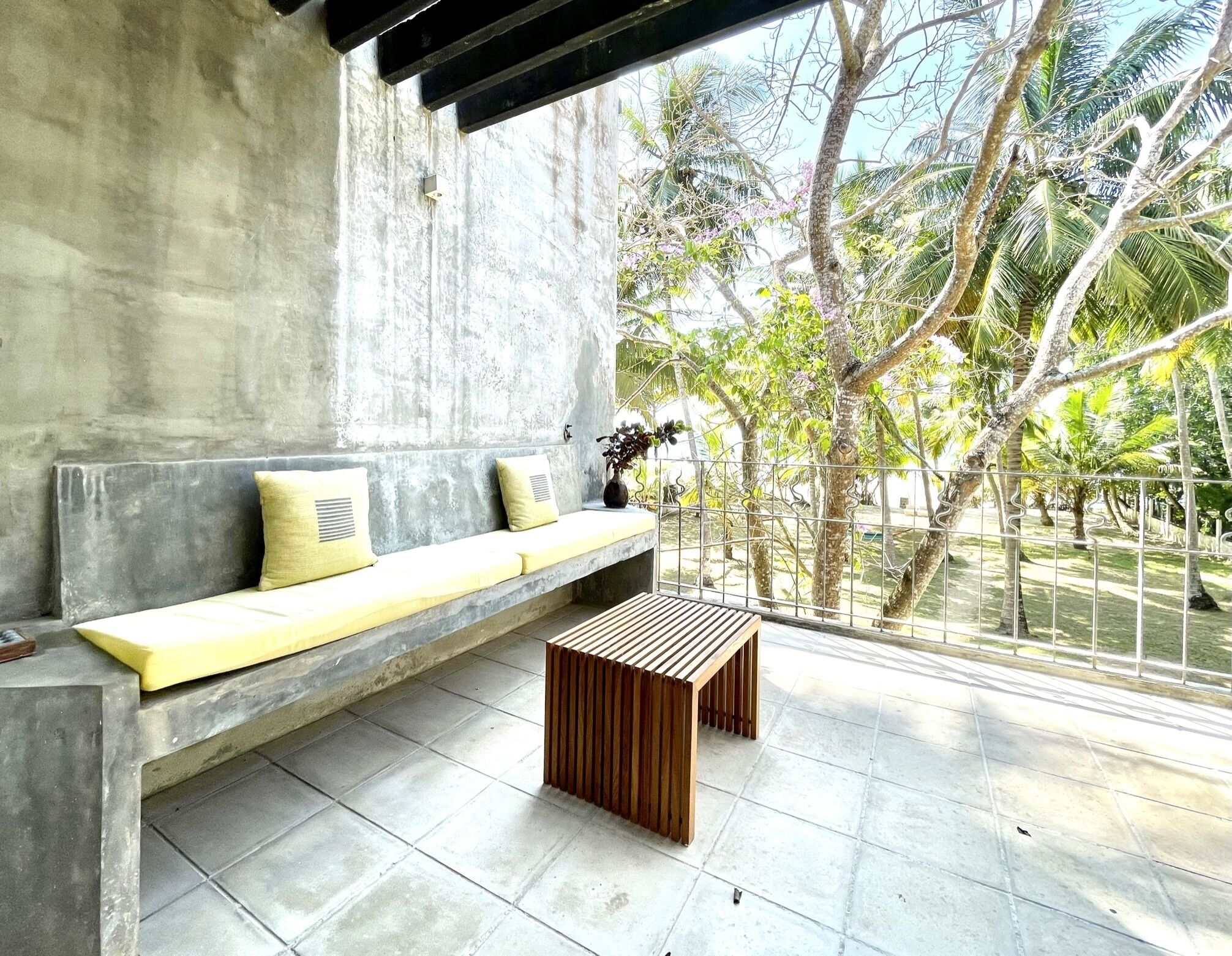 A modern outdoor seating area with a concrete bench and a wooden table, overlooking a garden with palm trees.