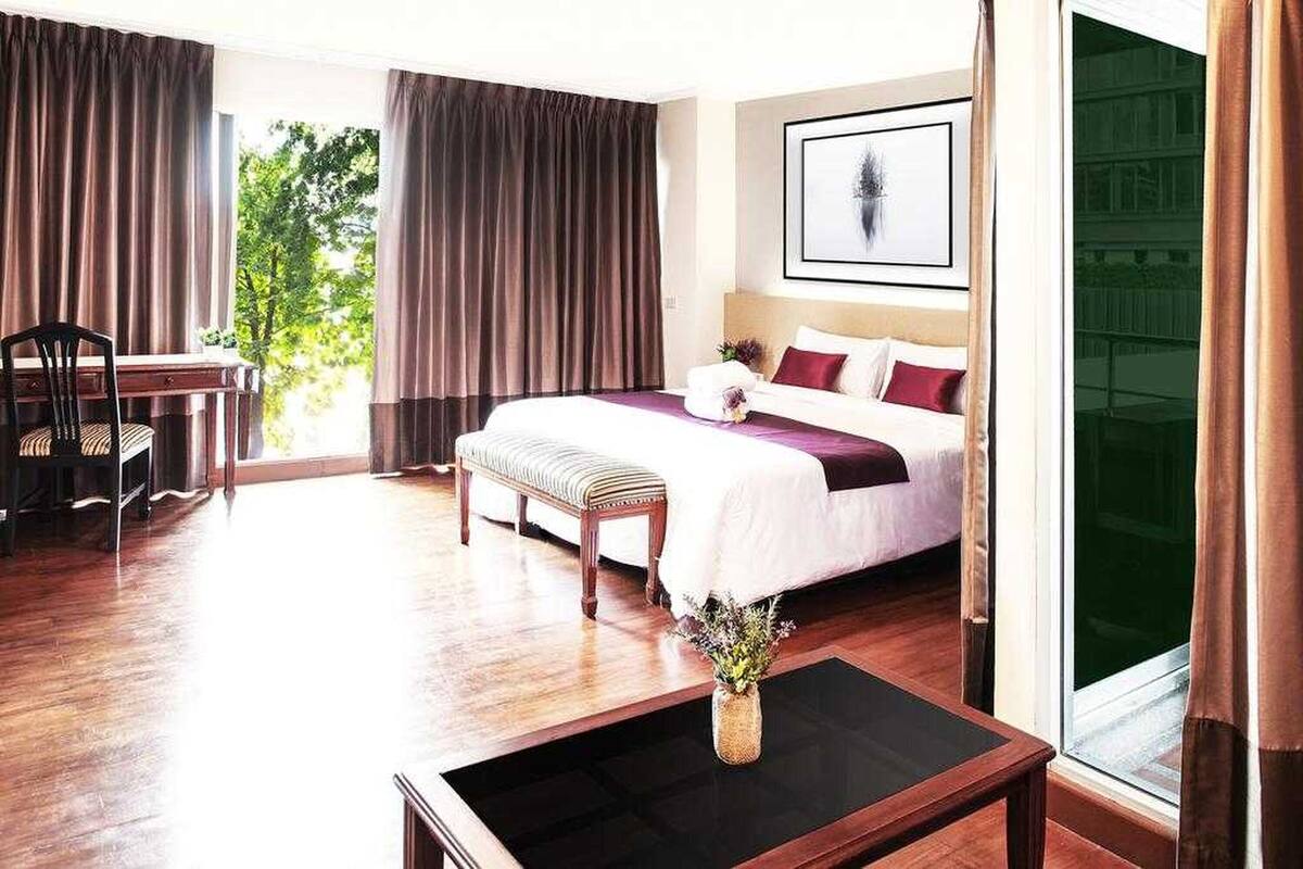 A hotel room with a large bed, a desk, and a chair. There is a window with curtains, a vase with flowers, and a framed picture on the wall.