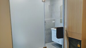 Ένα δωμάτιο ξενοδοχείου με μια γυάλινη πόρτα στο ντους, έναν λευκό νιπτήρα και ένα κρεβάτι με δύο πετσετέσ που είναι διπλωμένες σε ρολό.