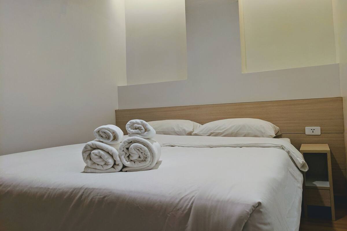 A neatly made bed with rolled towels, a wooden headboard, and a nightstand with a power outlet.