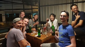 Μια ομάδα ανθρώπων κάθεται γύρω από ένα ξύλινο τραπέζι, μπροστά σε διάφορα μπουκάλια και ποτήρια, σε ένα δωμάτιο με μεγάλα παράθυρα και μεταλλική μπαλκονόπορτα.