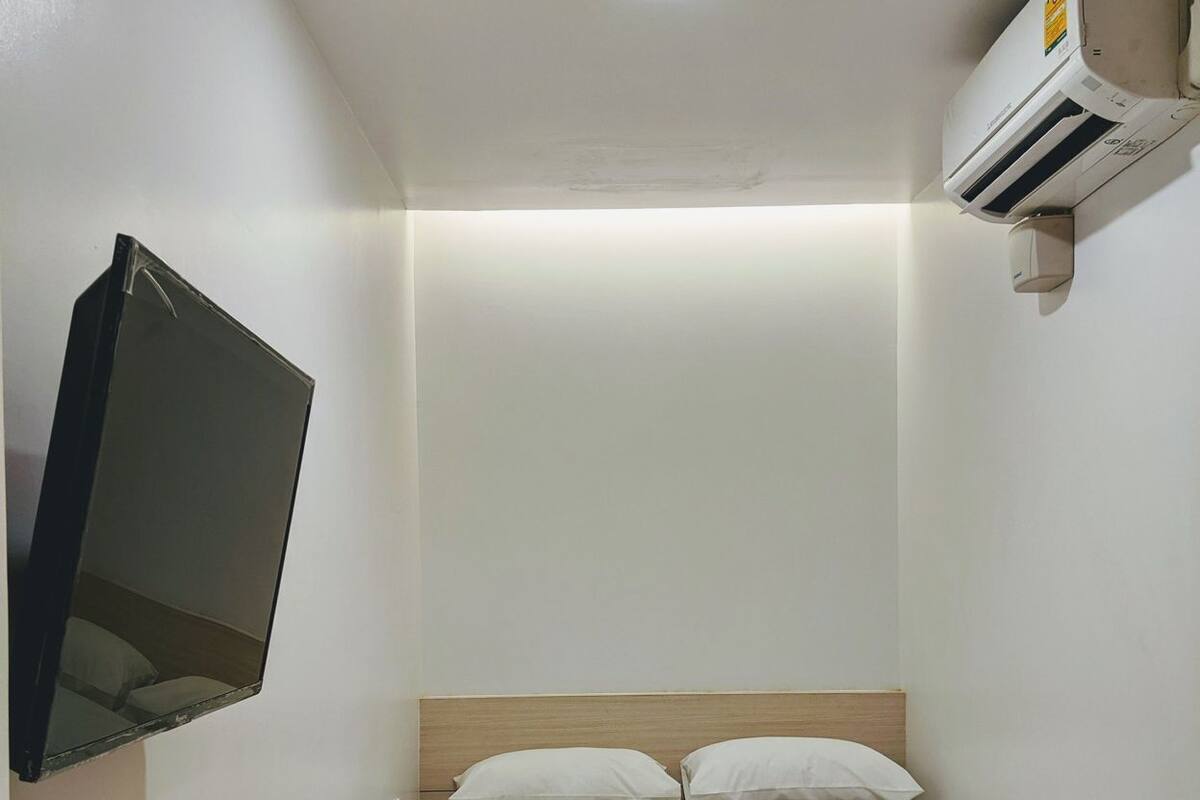 A single bed with white bedding, a mounted TV, an air conditioning unit, a wooden chair, and a remote control on a table.