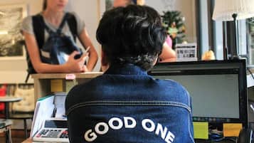 Una persona sentada en un escritorio con una computadora y un teléfono, usando una chaqueta de 'Good One Hostel & Cafe Bar'.