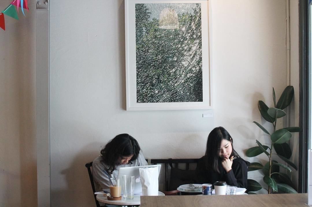 Two people seated at a table with a laptop, a glass, and a cup, in a room with a framed picture on the wall and a plant beside the window.