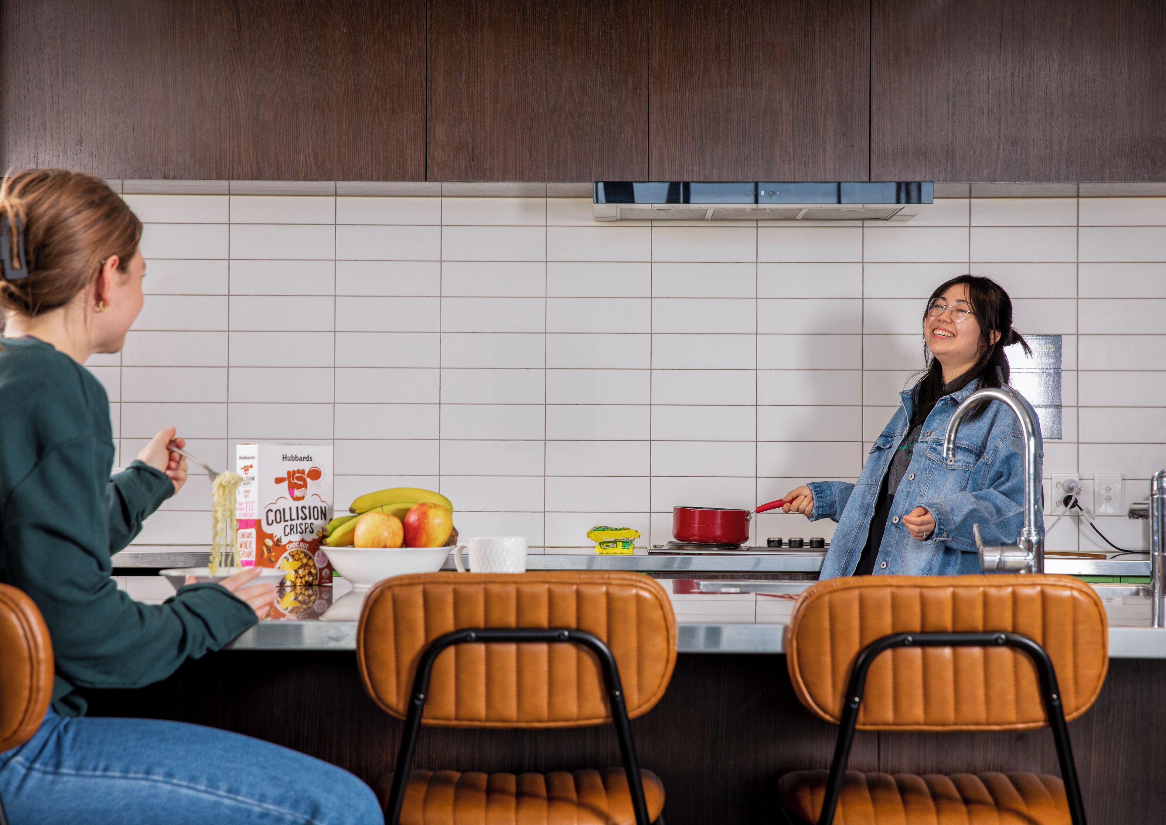 A kitchen with two people, one eating noodles and the other cooking. There are chairs, a table, and a fruit bowl with bananas and apples.