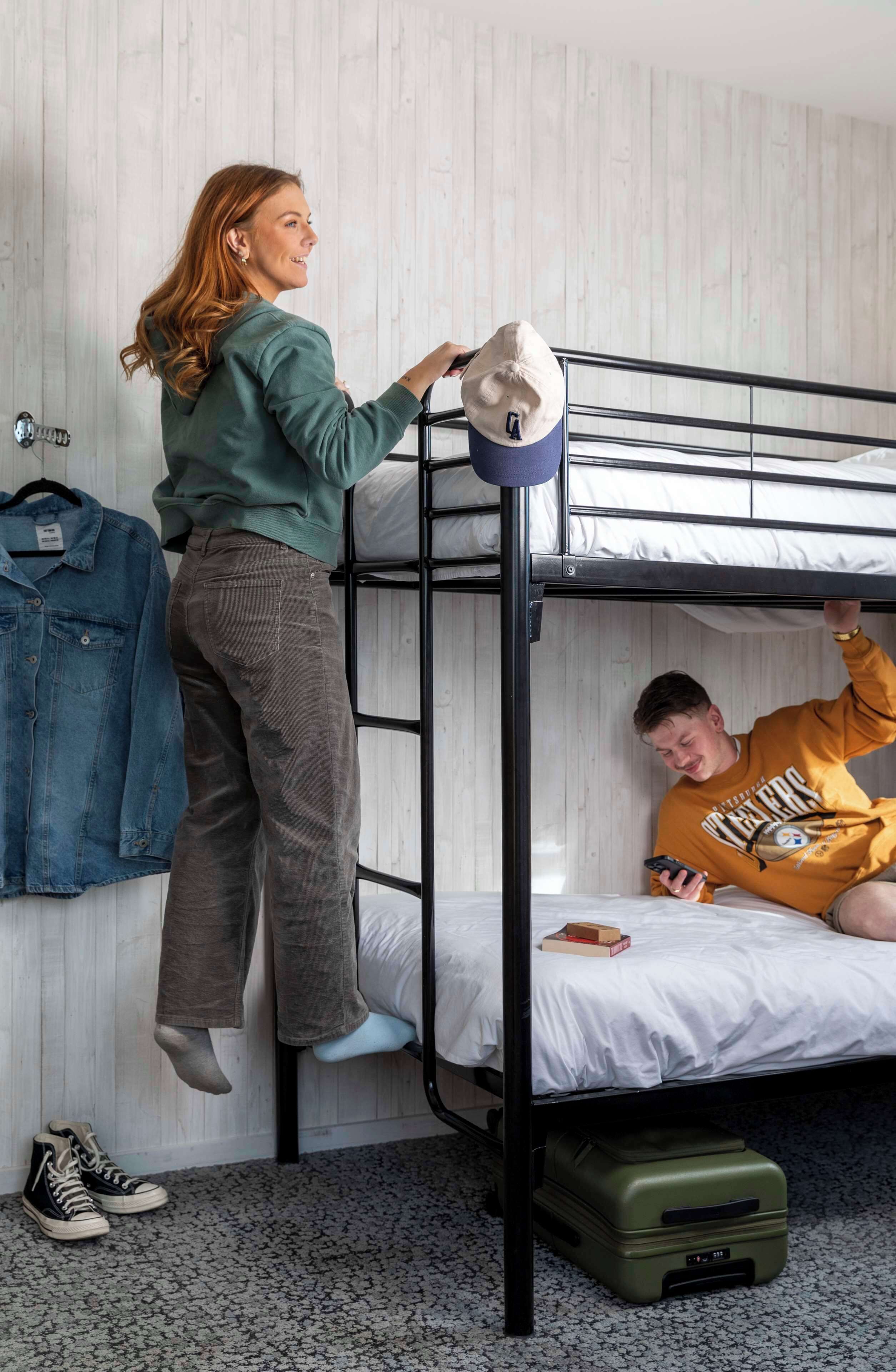 A bunk bed room with a person standing on the upper bunk and another person sitting on the lower bunk.