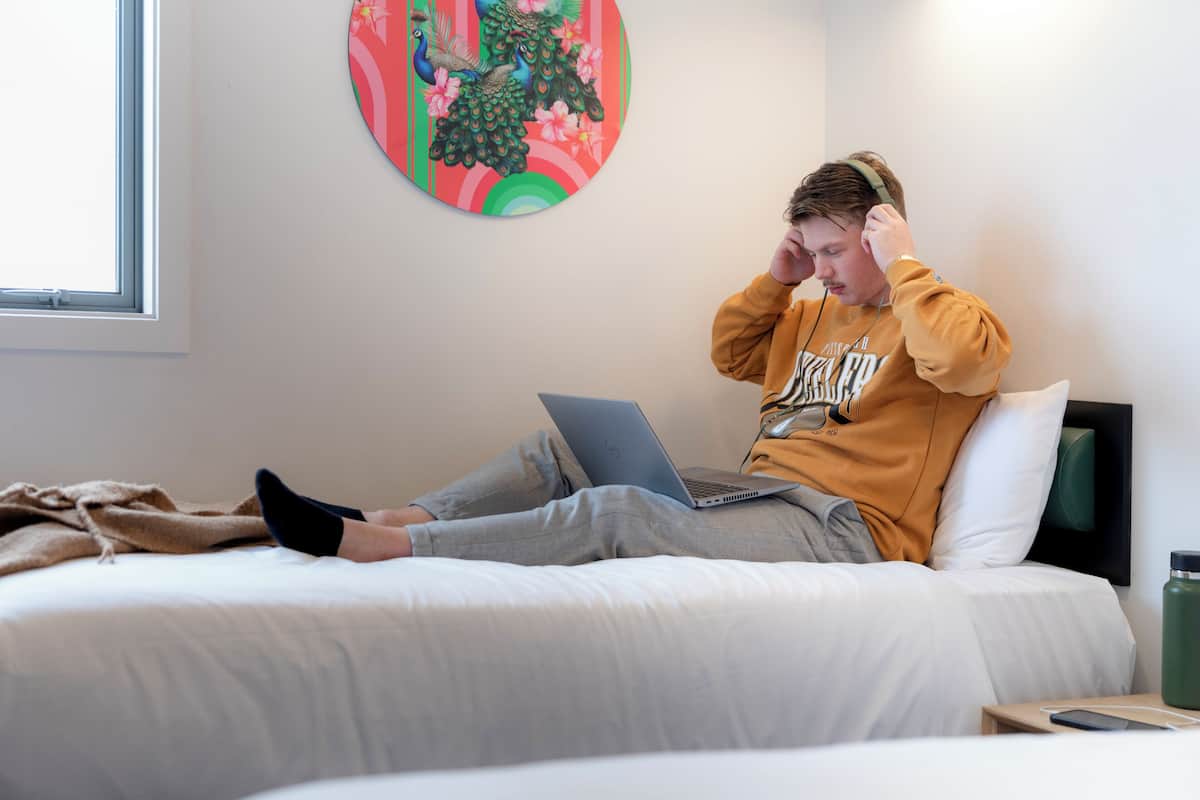 Una persona sentada en una cama con una computadora portátil, con sudadera con capucha, en una habitación con una obra de arte en la pared y una ventana.