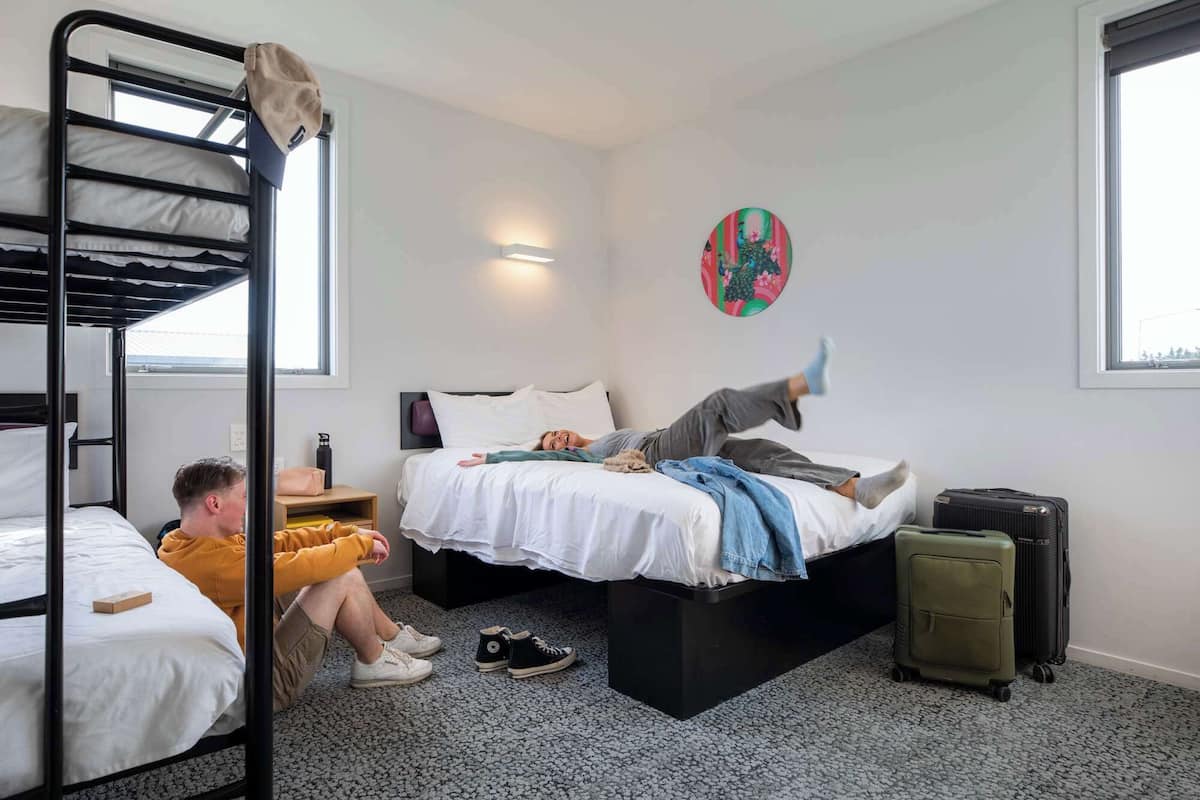 Un dormitorio con literas, una cama donde se ve a una persona acostada, una maleta y un reloj de pared.