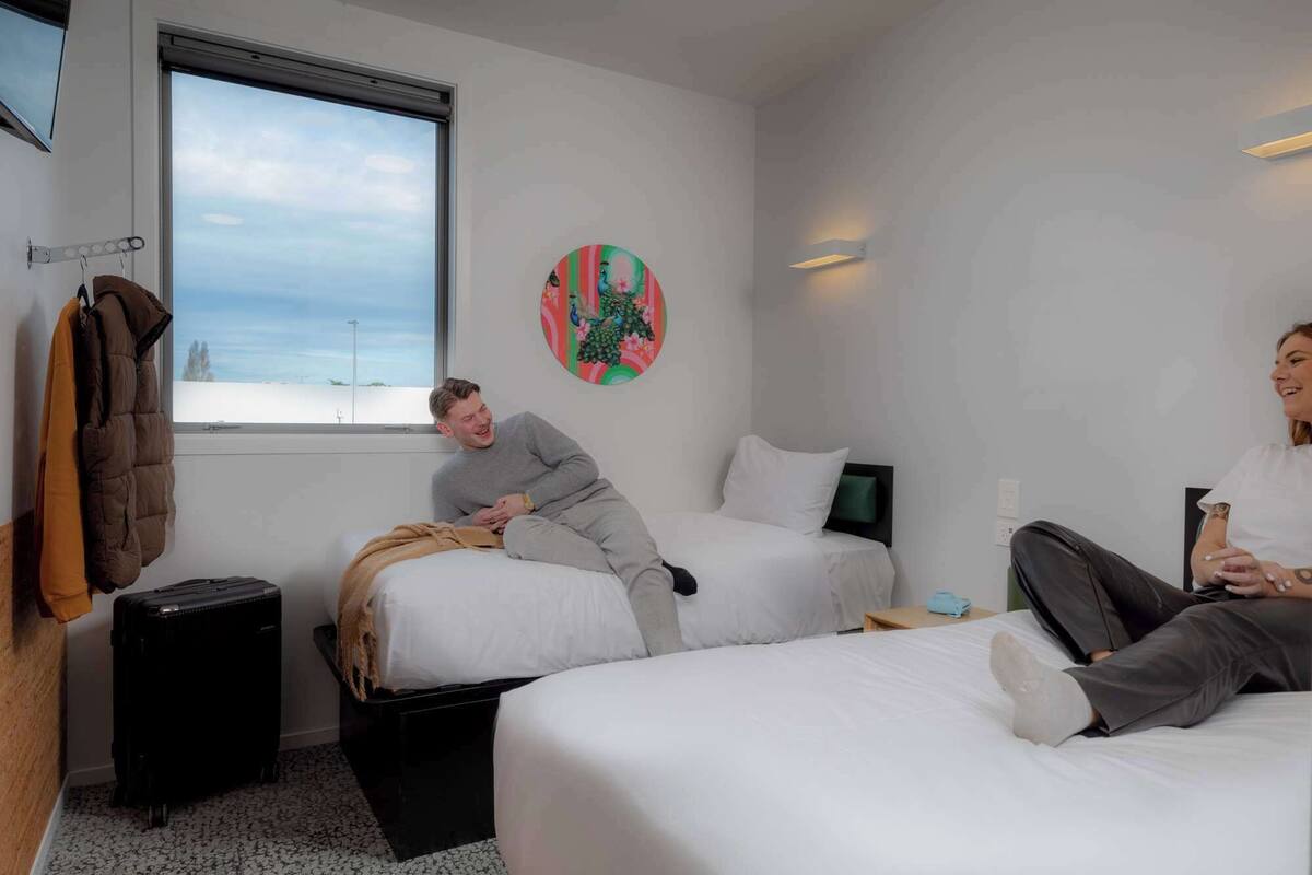 Habitación de hotel con dos camas, ventana con vistas, un adorno en la pared y una maleta en el suelo.