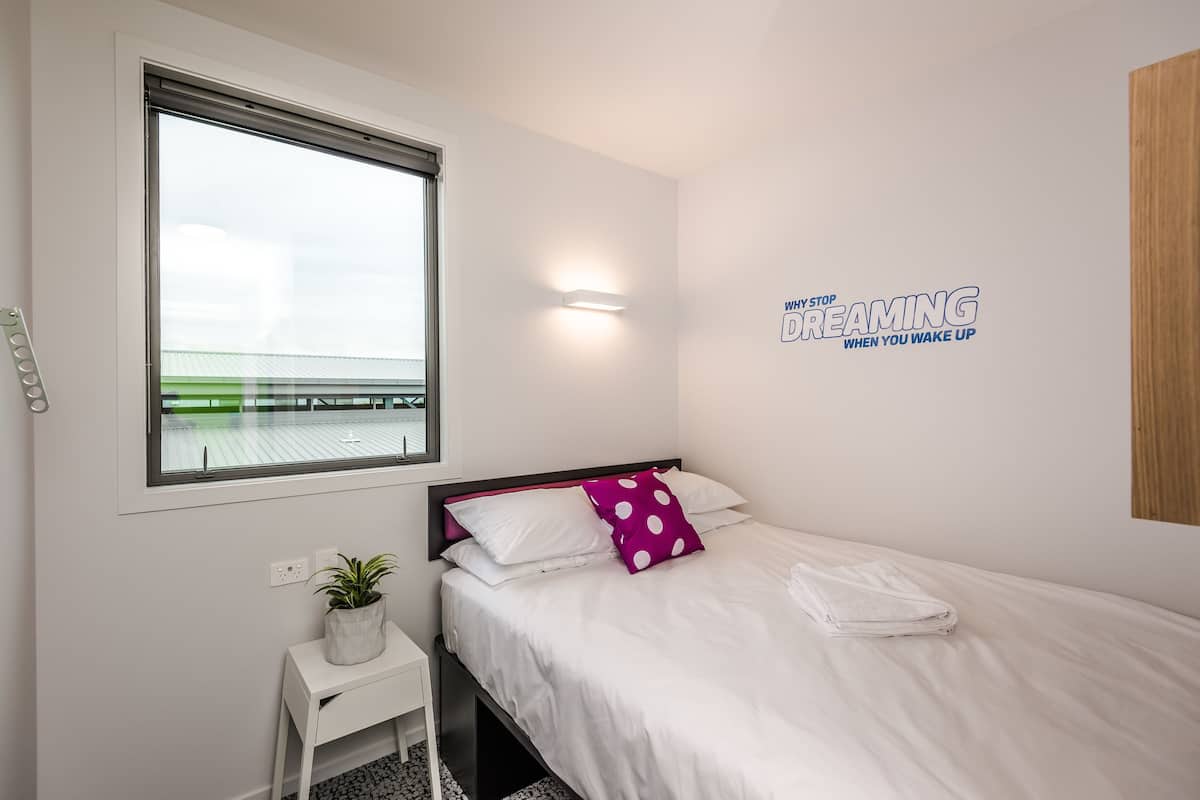 Un dormitorio con una cama, una mesita de noche con una planta, una ventana y una pared con una cita motivacional.
