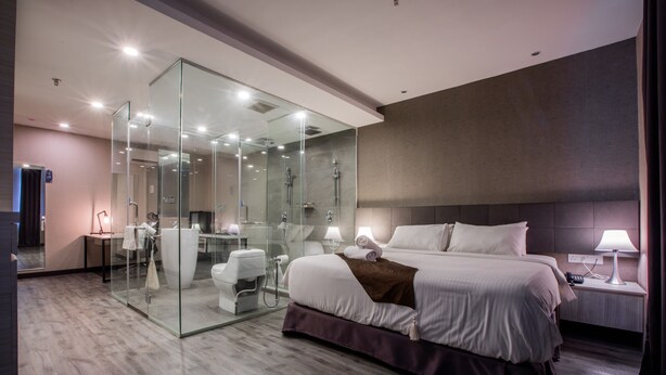 Ein modernes Hotelzimmer mit einer gläsernen Dusche, einem großen Bett, einem Nachttisch und einem Beistelltischchen mit einer Lampe.