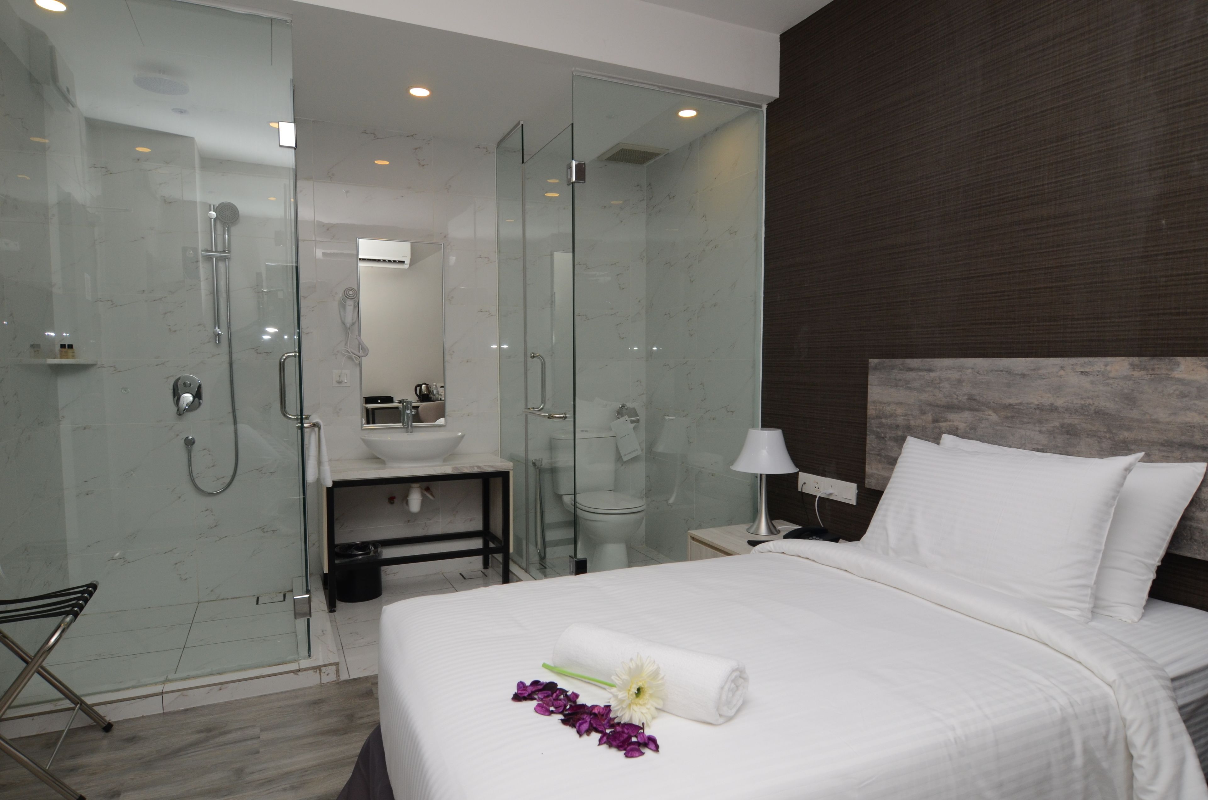 Ein modernes Hotelzimmer mit einer gläsernen Dusche, einem Badezimmer mit Waschbecken und Toilette, einem ordentlich bezogenen Bett mit einem gerollten Handtuch und Blumen sowie einem dunklen Holz-Kopfteil.