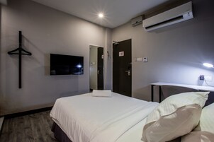 A hotel room with a bed, a flat-screen TV mounted on the wall, an air conditioning unit, a coat rack, and a desk with a chair.
