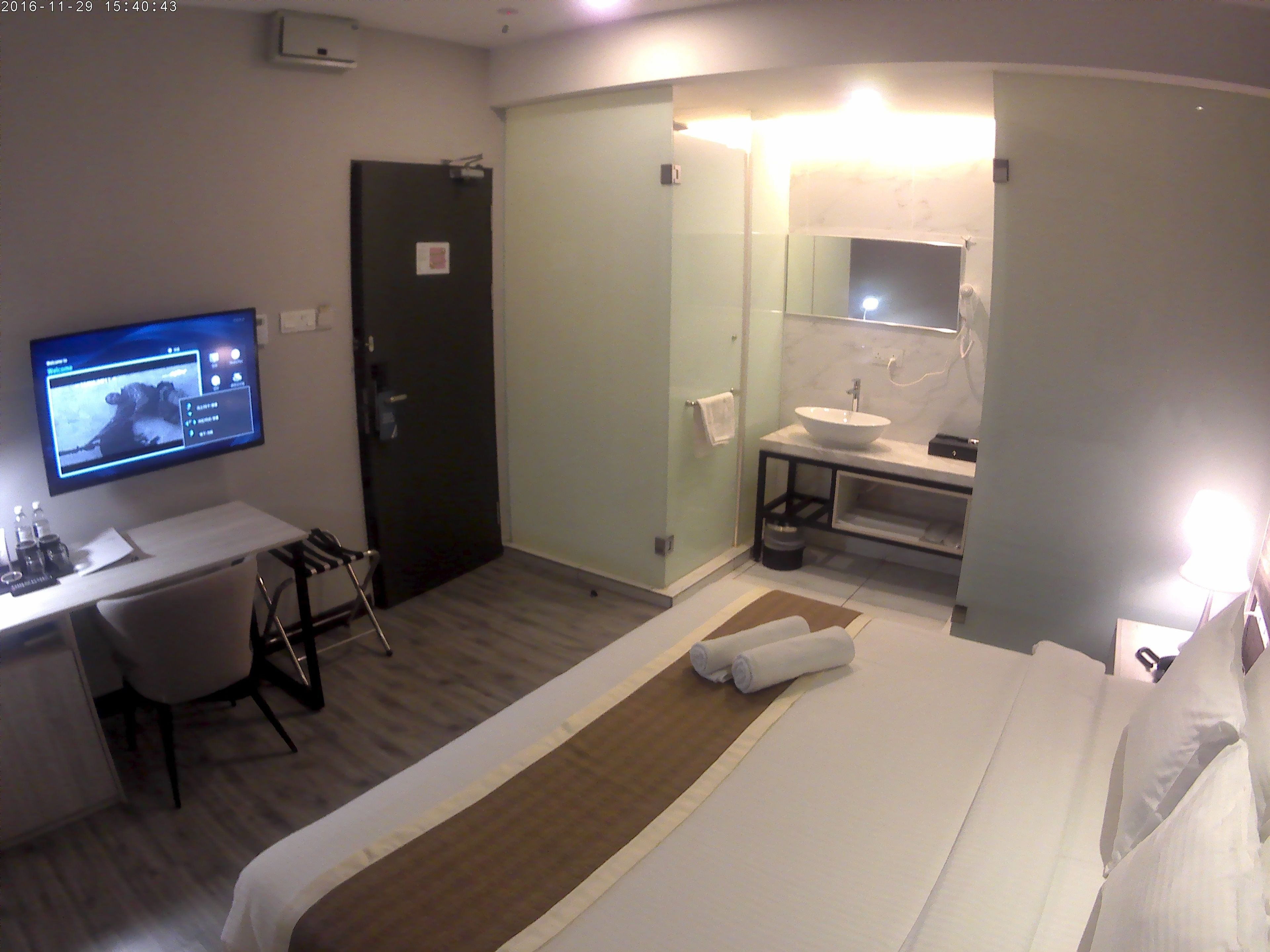 A hotel room with a bed, a desk with a computer, a TV, a bathroom with a sink and mirror, and a door.