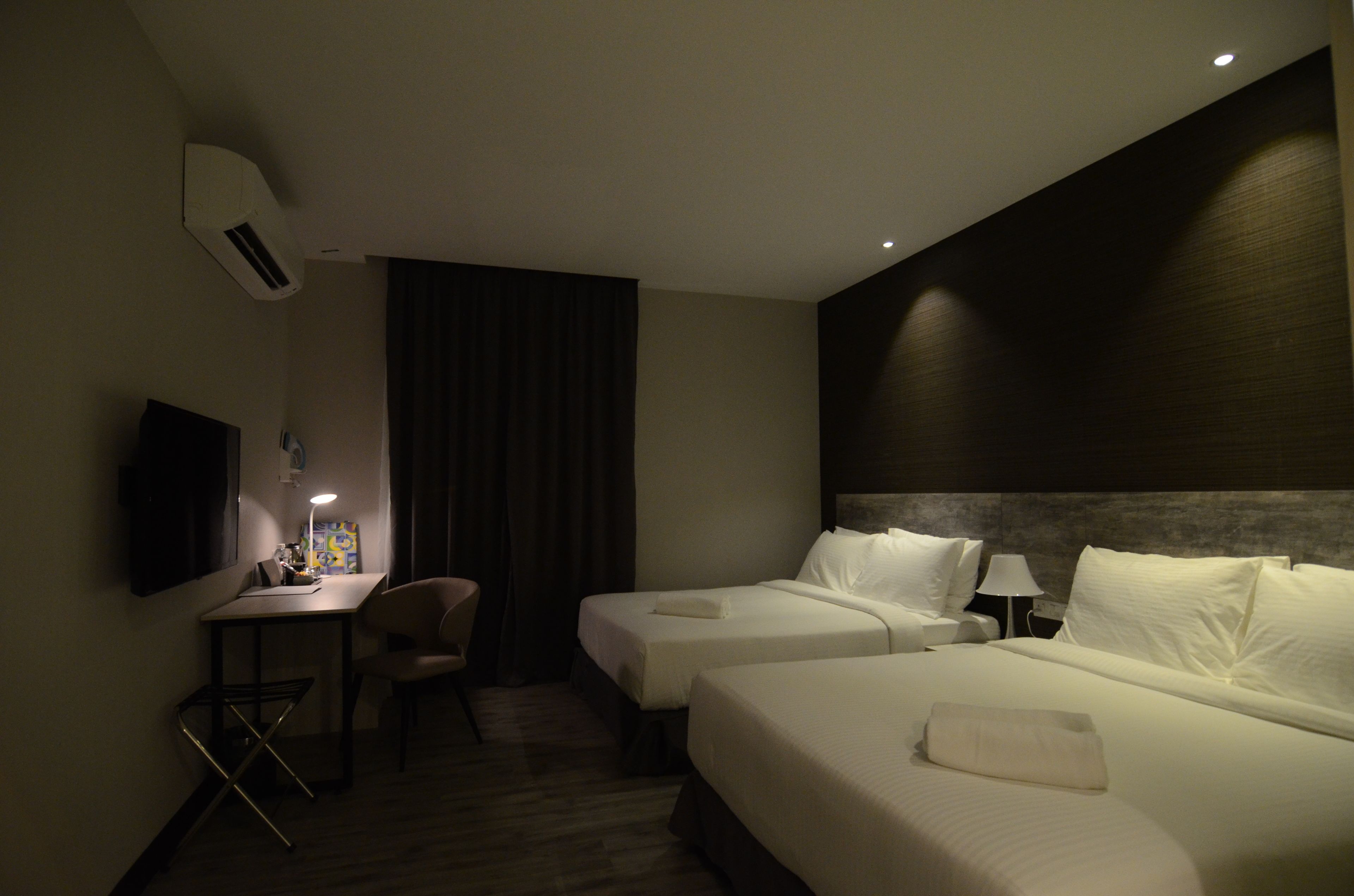 A hotel room with two beds, a desk, a chair, a TV, and an air conditioning unit.