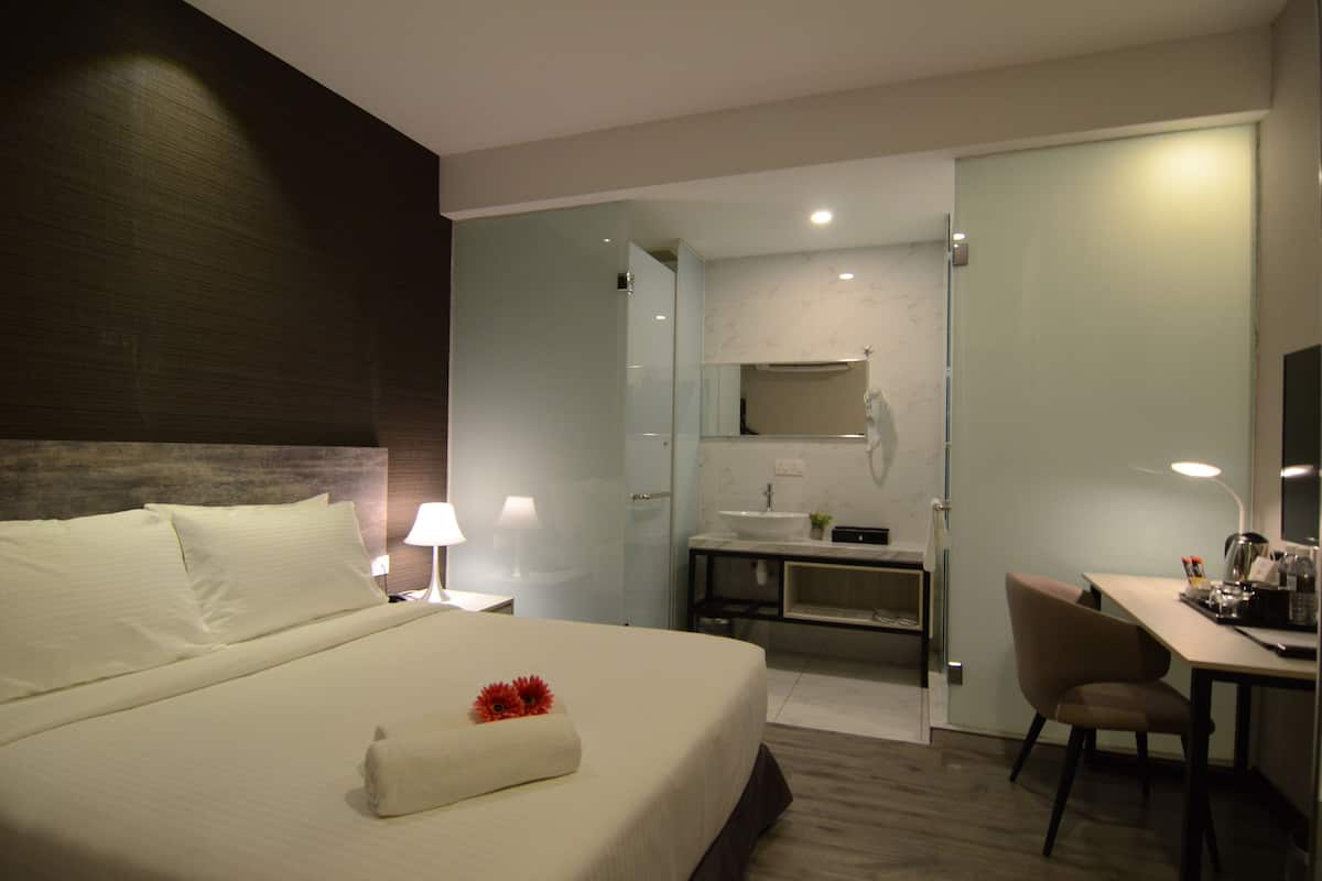 Una habitación de hotel moderna con una cama grande, un escritorio, una silla, un baño con lavamanos y espejo, y zona de ducha.