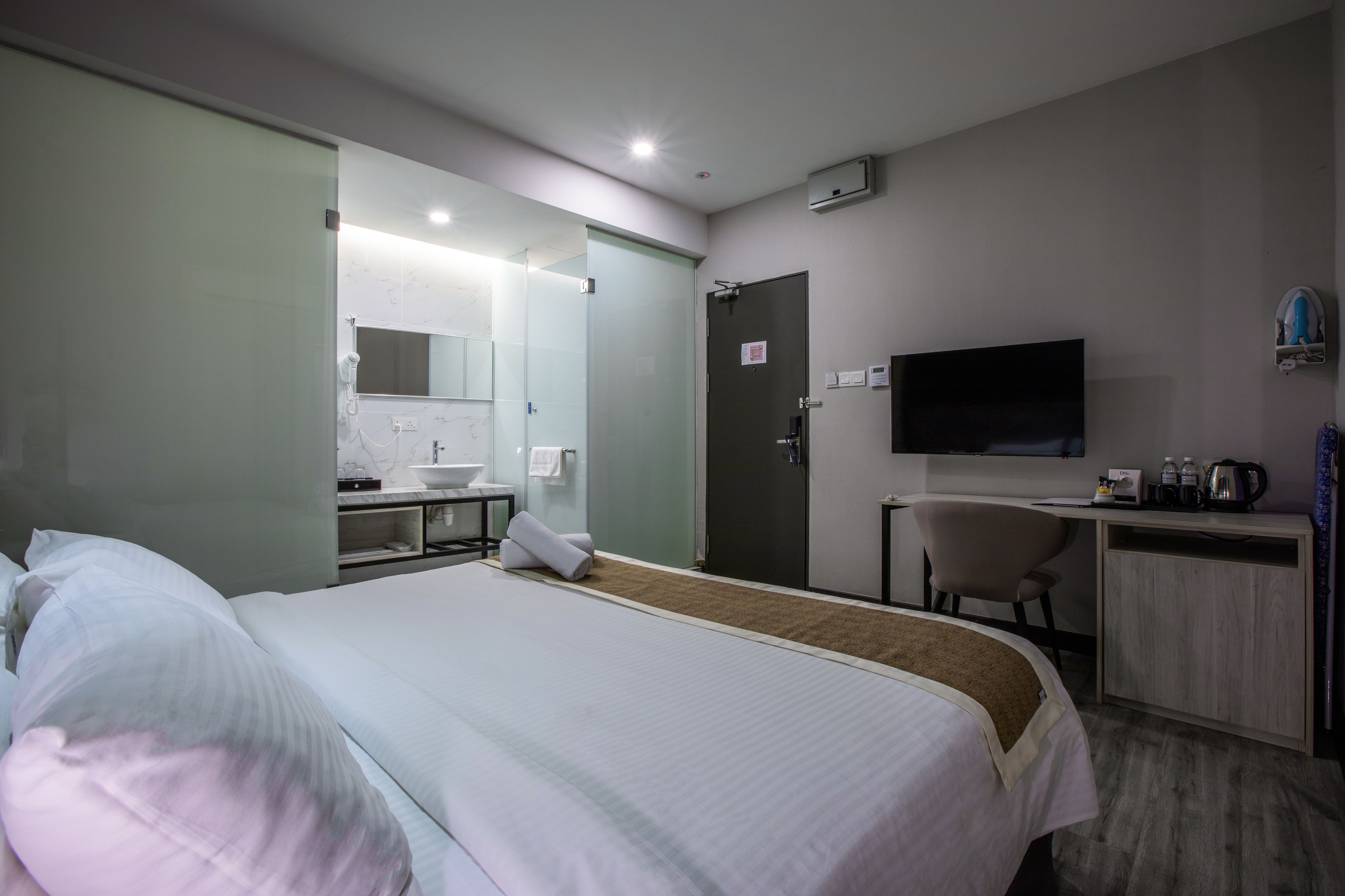 Habitación de hotel con cama, escritorio, televisor, espejo y baño con lavamanos y toallero.