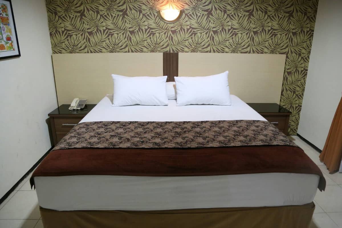 Una cama bien tendida, con almohadas blancas y una manta marrón, un teléfono en la mesita de noche y una pared con un diseño de hojas.