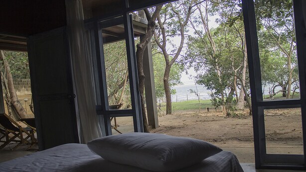 A bed with a pillow in a room with large windows overlooking a dirt area with trees and a body of water. - Wapi Bungalowi Yala (Thissamaharama)