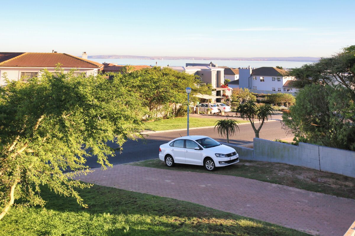 A residential street with houses, a white car parked on the side, and a view of the sea in the background.