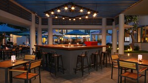 An outdoor dining area with a central bar, multiple tables and chairs, umbrellas, and a ceiling with hanging lights. - ÊMM Hotel Hoi An (Da Nang)