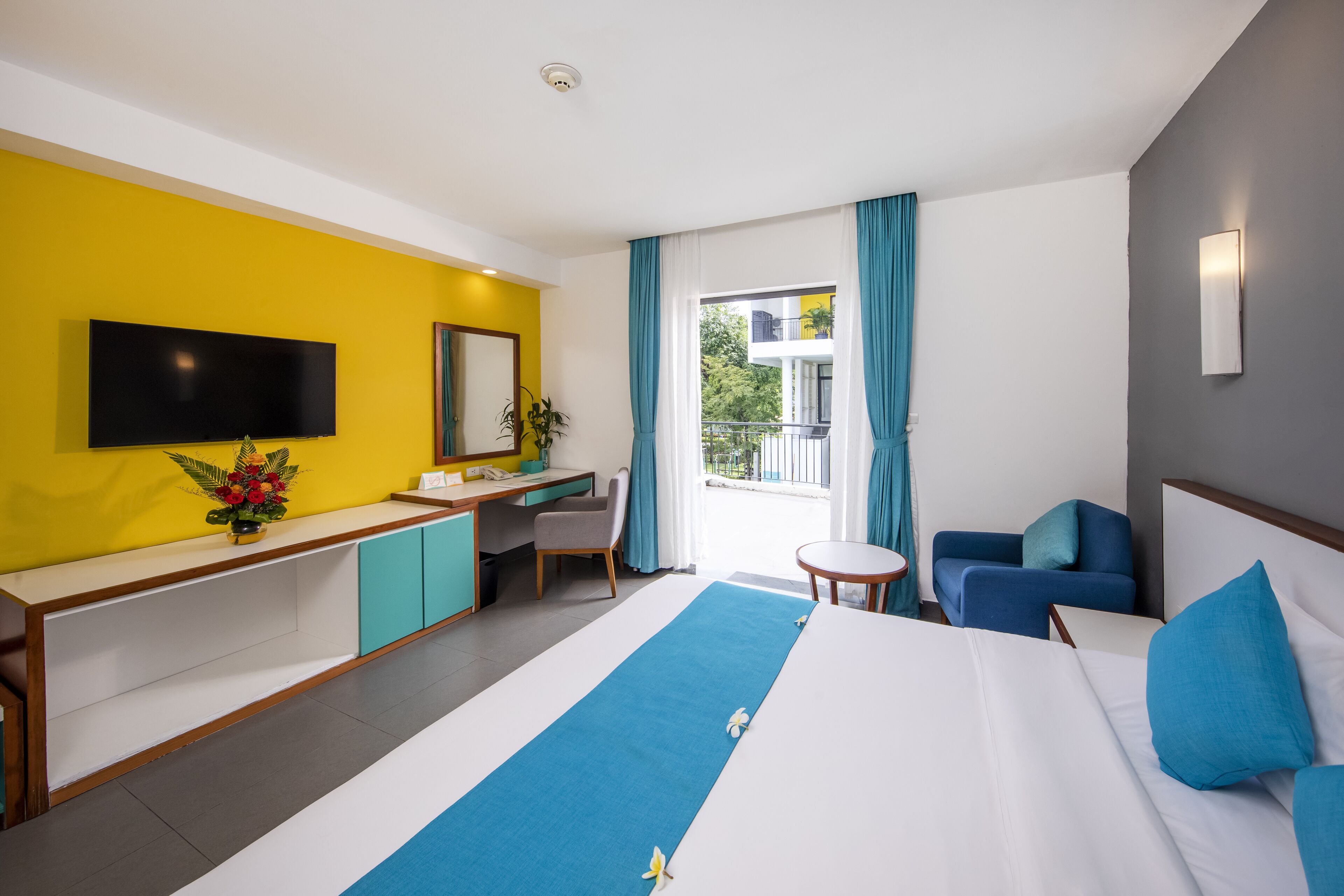A hotel room with a bed, a desk, a chair, a TV, and a balcony with curtains.