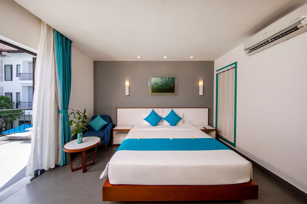 A hotel room with a bed, a small table, a chair, a potted plant, and a view of the outside.