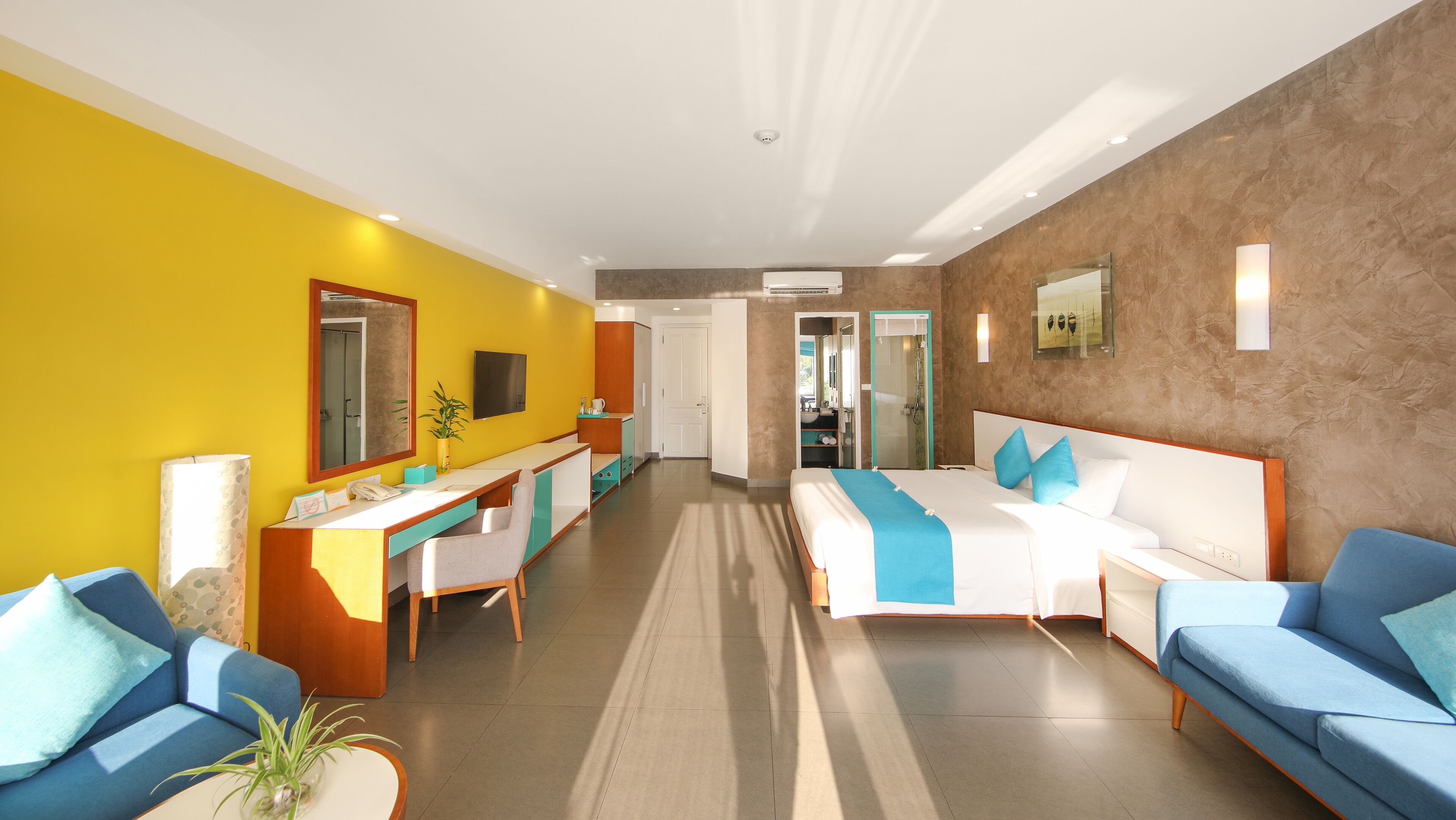 Habitación de hotel con cama, dos sofás azules, un escritorio con silla, un espejo y un baño visible a través de una puerta abierta.
