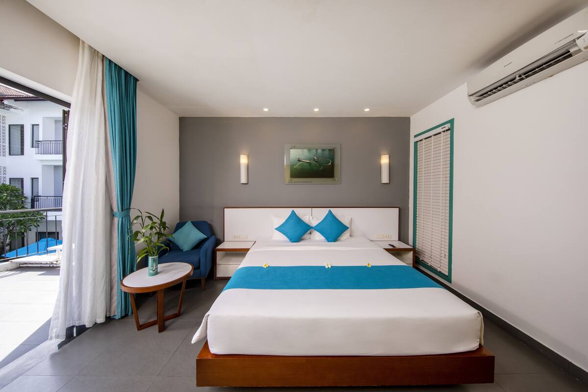 A hotel room with a large bed, a bedside table, a chair, a small table with a plant, and a view of the outside.