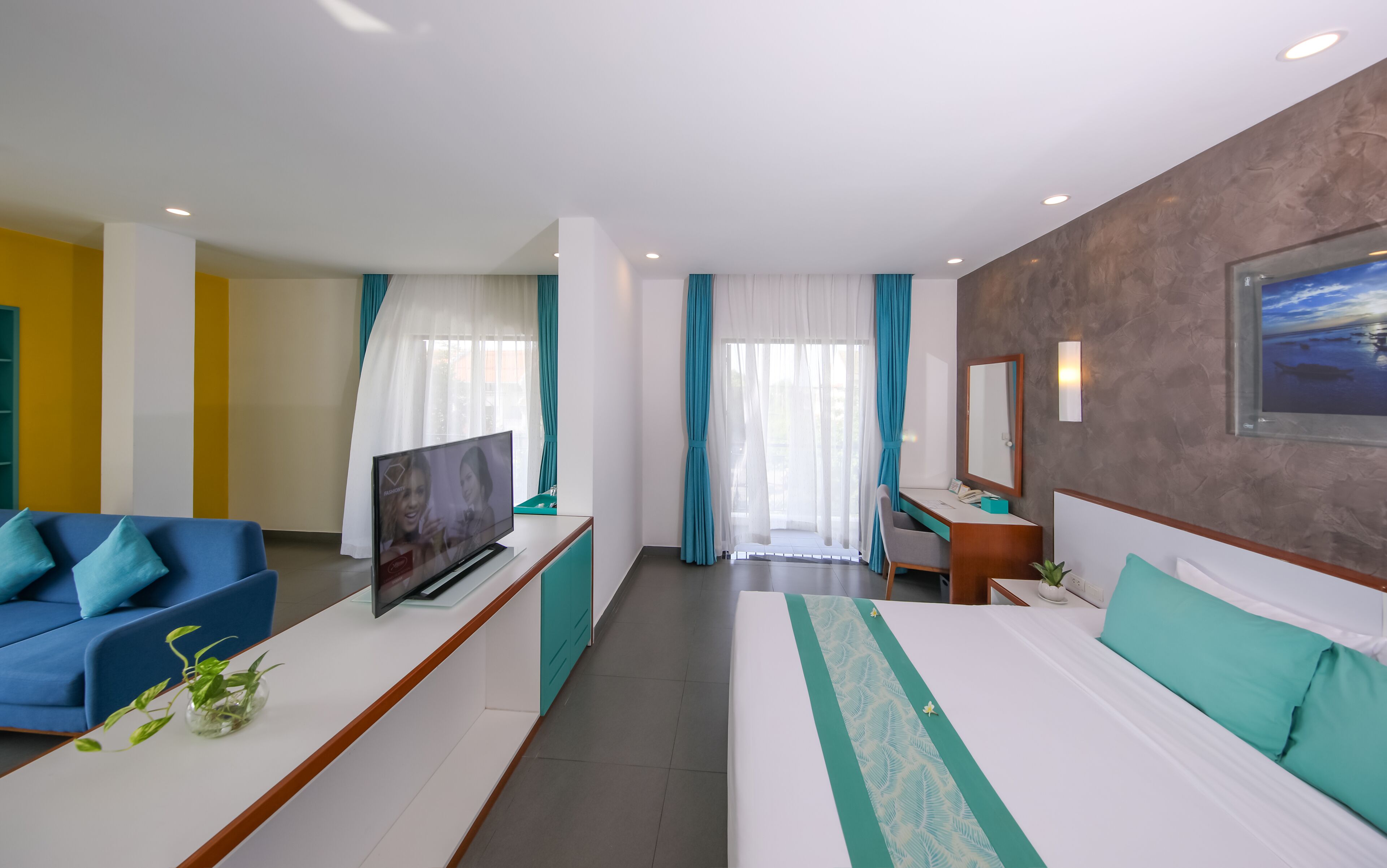 A modern hotel room with a large bed, a desk, a TV, and a seating area with a blue sofa.