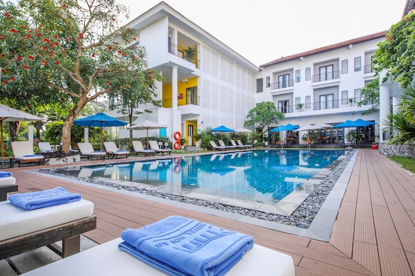 A swimming pool area with lounge chairs and umbrellas, surrounded by a building with balconies and a poolside deck. - ÊMM Hotel Hoi An (Da Nang)