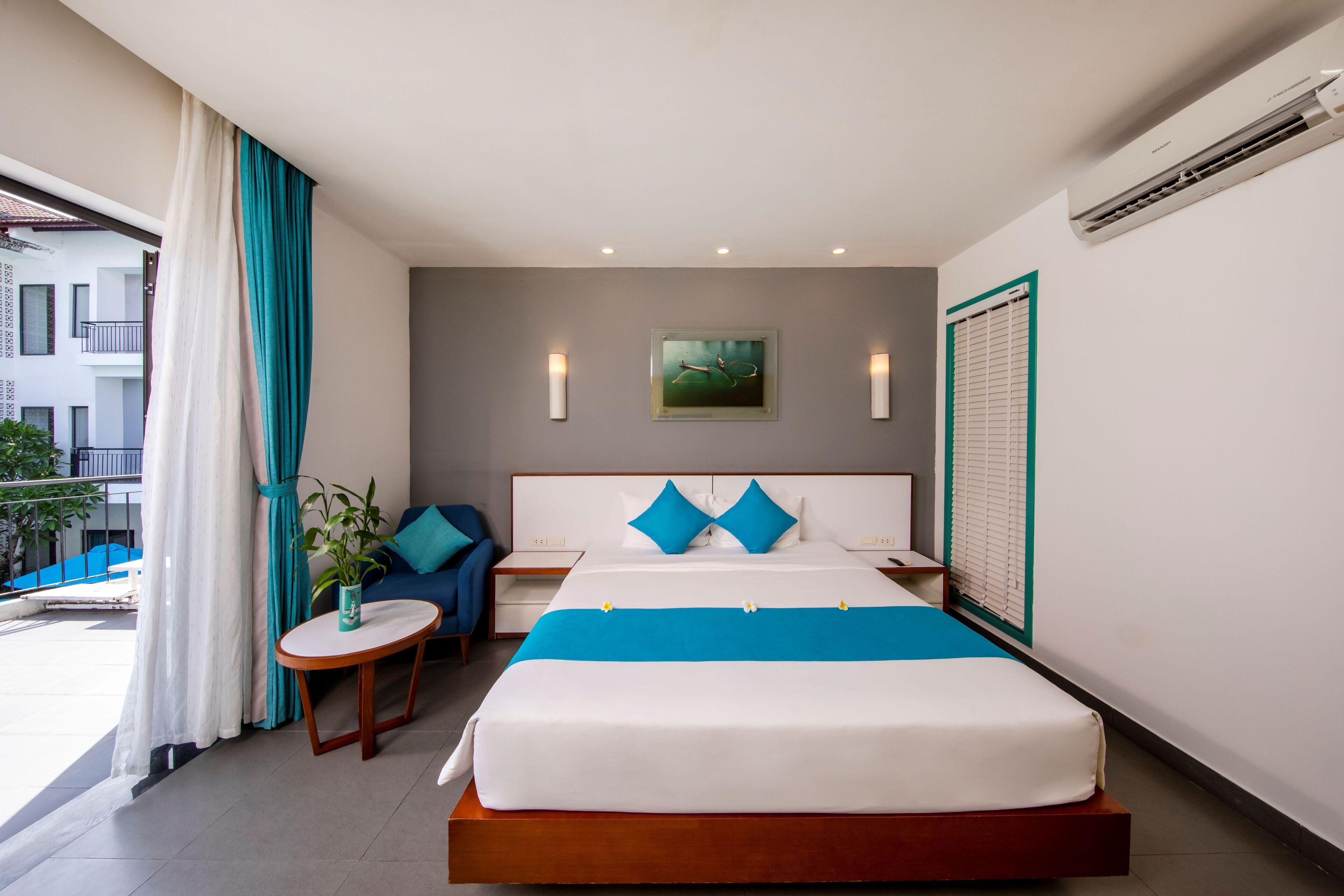 A hotel room with a bed, a small table, a chair, a potted plant, and a view of the outside.