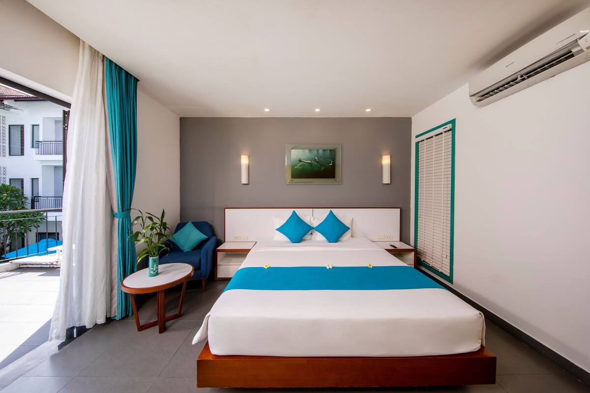 A hotel room with a bed, a small table, a chair, a potted plant, and a view of the outside.
