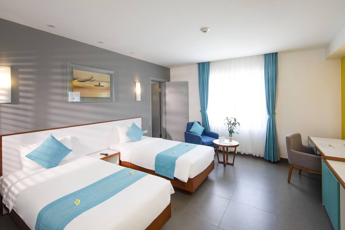 A hotel room with two beds, a desk, a chair, a small table, and a window with curtains.
