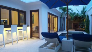 An outdoor seating area with white chairs and a blue umbrella, a pool, and a glass door leading to a dining area. - Kamil Villas (Seminyak)