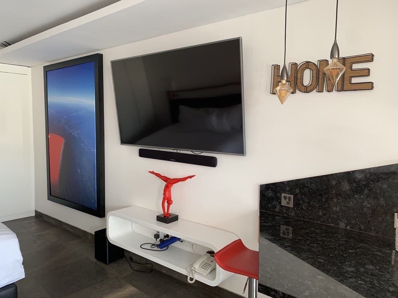 Una habitación de hotel moderna con un televisor de pantalla plana grande montado en la pared, una escultura roja sobre una consola blanca, y un cartel que dice 'HOME'.