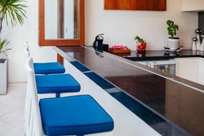 A modern kitchen with a long countertop, blue cushioned seating, and various kitchen items. - Kamil Villas (Seminyak)