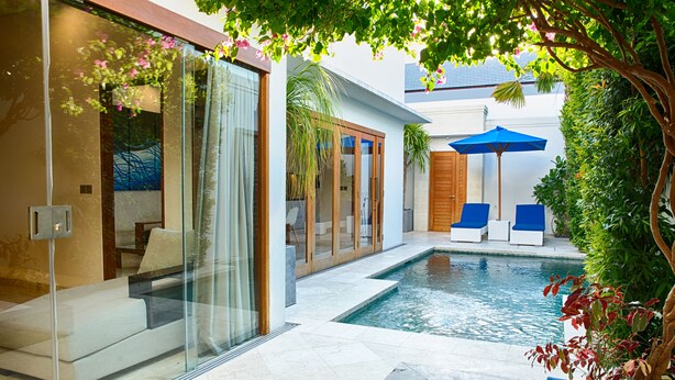 A modern outdoor pool area with glass walls, a lounge with blue chairs, and a wooden deck. - Kamil Villas (Seminyak)