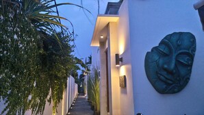 A narrow corridor with a white wall on the right, a decorative face sculpture, and a row of potted plants on the left. - Kamil Villas (Seminyak)