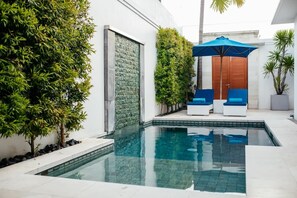 A modern outdoor pool area with a clear pool, two blue lounge chairs, and a wooden door. - Kamil Villas (Seminyak)