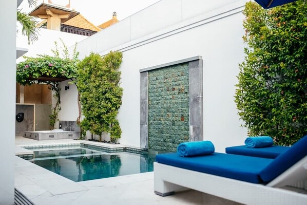 A modern indoor pool area with a clear pool, blue lounge chairs, and lush greenery. - Kamil Villas (Seminyak)
