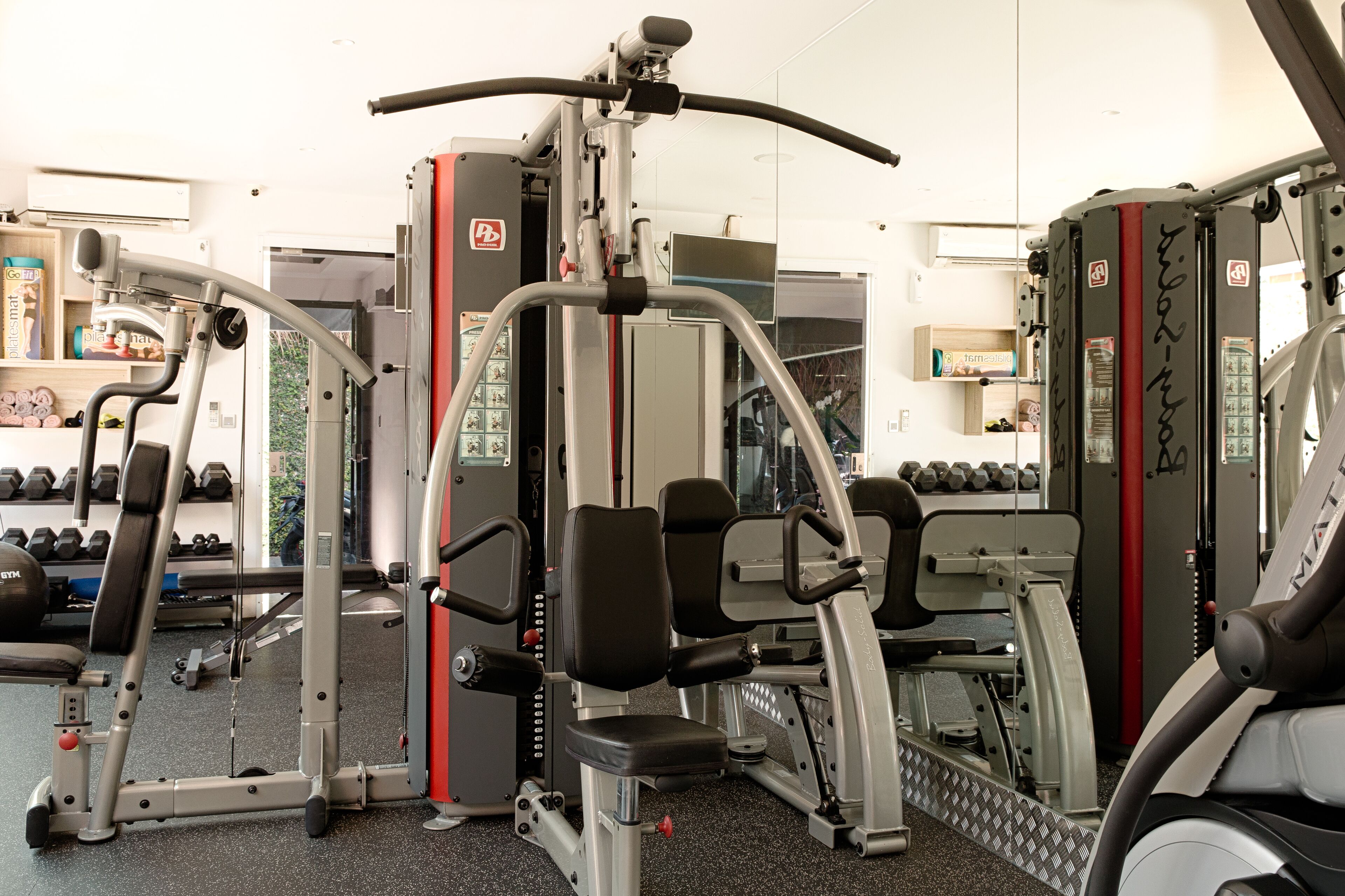 Ein Fitnessstudio mit verschiedenen Trainingsgeräten, einer Spiegelwand und Hanteln auf Regalen.