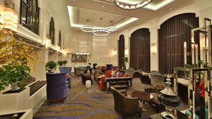 A spacious lobby with a variety of seating options, including sofas and armchairs, arranged around a central coffee table. There are multiple decorative plants and a large mirror reflecting the room.