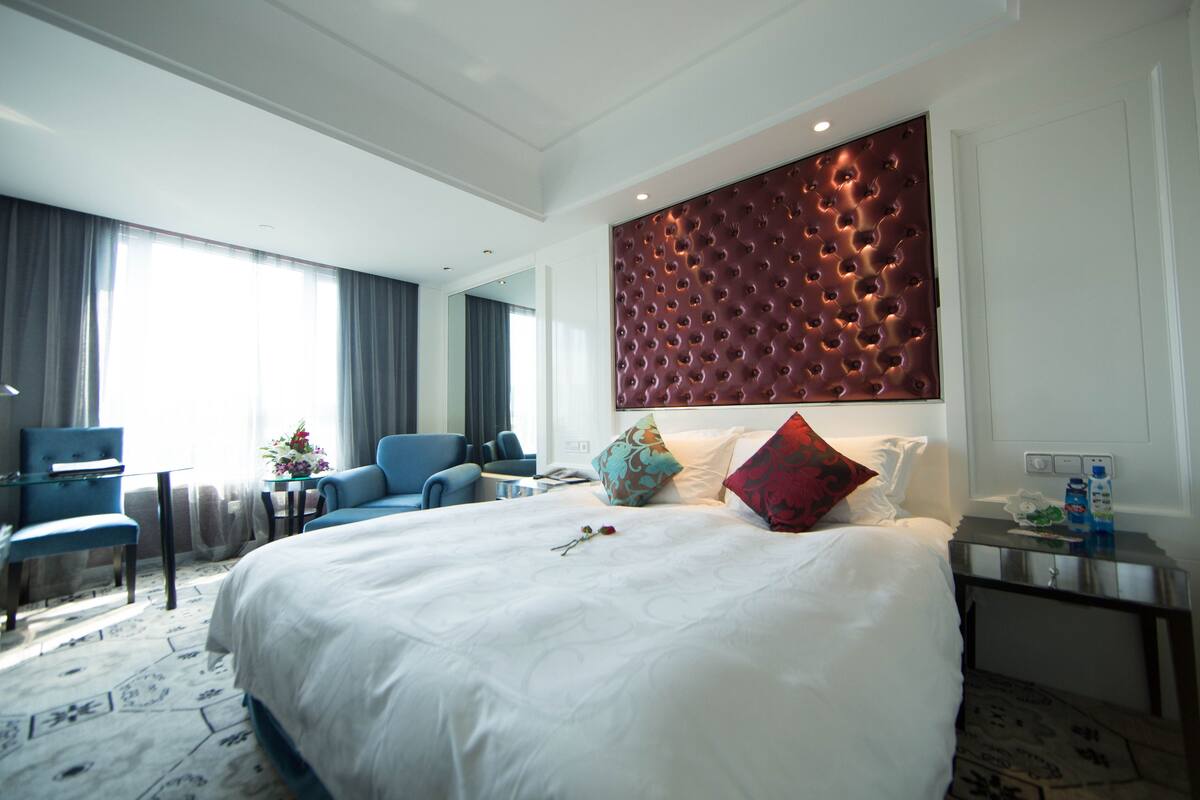 Habitación de hotel con una cama grande, cabecero capitonado, zona de estar con un sillón azul, una mesita con un jarrón de flores y una mesita de noche con una botella de agua.